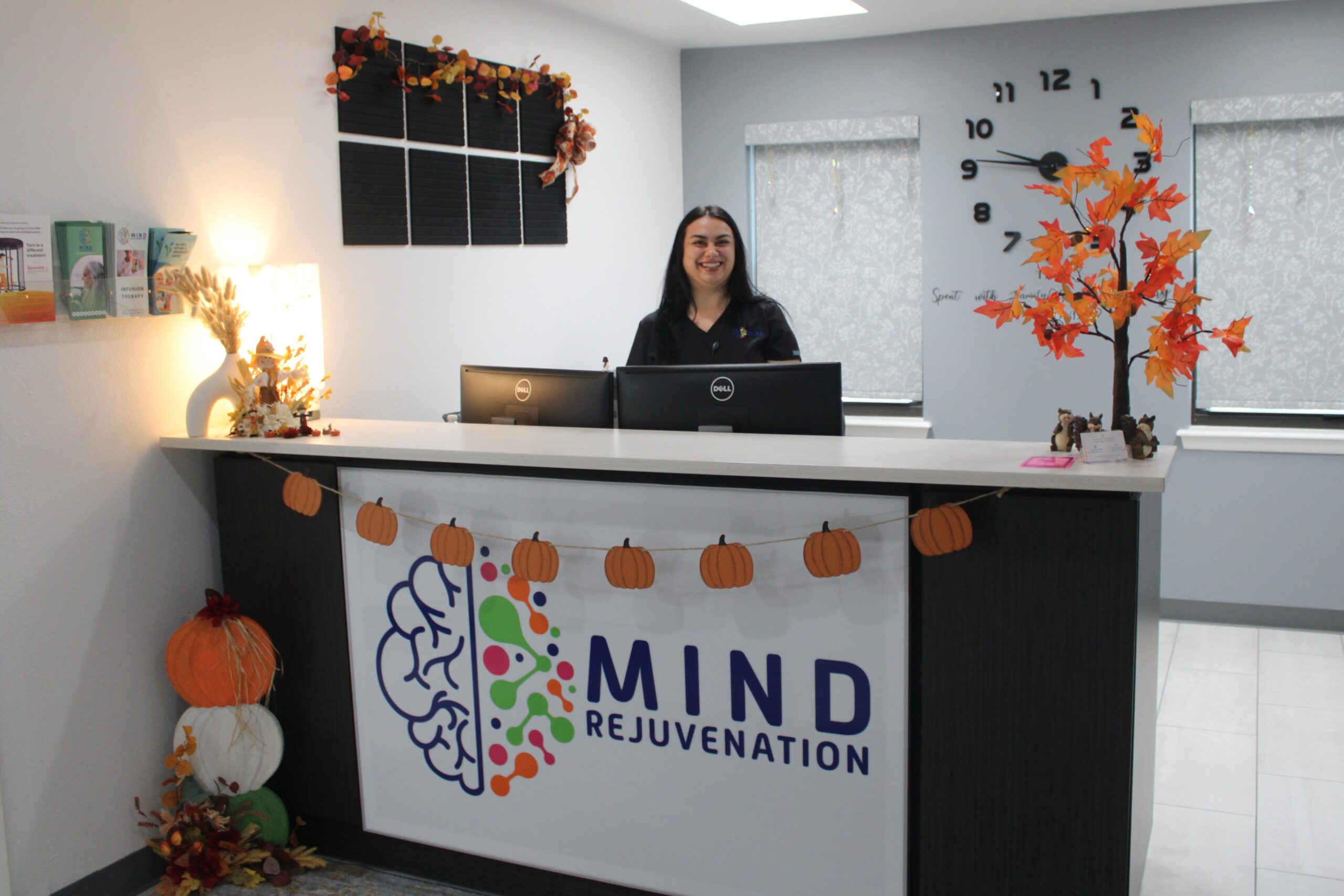 Smiling receptionist behind the Mind Rejuvenation front desk, surrounded by autumn-themed decor and wellness branding. Featured on the Home page to reflect warmth, professionalism, and a welcoming check-in experience for mental health care.