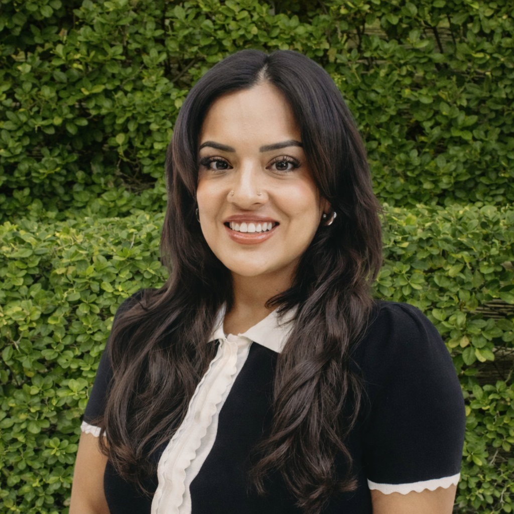 Board-Certified Psychiatric Mental Health Nurse Practitioner, Mical Pacheco, MSN, APRN, PMHNP-BC, smiling in a black shirt with white collar outdoors against a green hedge. Featured on the Meet Our Team page to reflect compassionate care, clinical expertise, and a welcoming presence at Mind Rejuvenation.