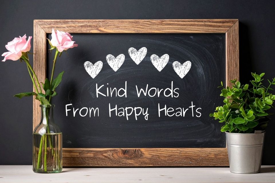 A chalkboard with five hand‑drawn white hearts above the phrase “Kind Words From Happy Hearts,” displayed between a vase of pink roses and a small potted plant.