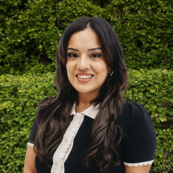 Board-Certified Psychiatric Mental Health Nurse Practitioner, Mical Pacheco, MSN, APRN, PMHNP-BC, smiling in a black shirt with white collar outdoors against a green hedge. Featured on the Meet Our Team page to reflect compassionate care, clinical expertise, and a welcoming presence at Mind Rejuvenation.